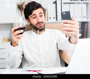 Happy young office worker with glass of wine taking selfie on his smartphone at modern office Stock Photo