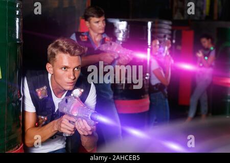 Young positive cheerful glad guy holding colored laser guns and took aim during laser tag game in labyrinth Stock Photo