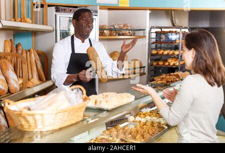 Polite male baker working behind counter in store of his bakery, serving female customer Stock Photo