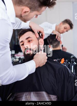 Pleasant  male hairdresser accurately cutting beard of client at hair salon Stock Photo