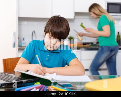 Positive schoolboy doing homework using laptop at kitchen interior, woman cooking at background Stock Photo