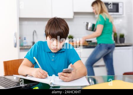 Positive schoolboy doing homework using laptop at kitchen interior, woman cooking at background Stock Photo