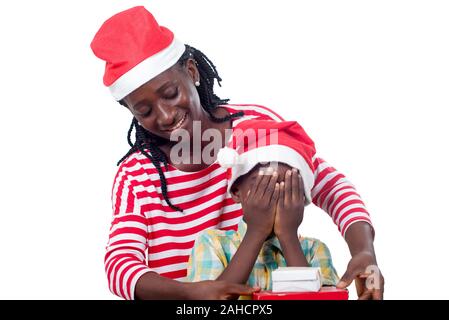 smiling young woman in Santa Claus hat holds a gift in front of her child in Santa Claus hat and eyes closed by her hands. Stock Photo