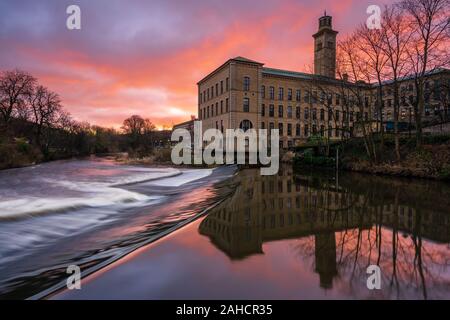 A vibrant sunrise frames New Mill at the Saltaire UNESCO World Heritage Site, with the distinctive architecture reflected in the River Aire. Stock Photo