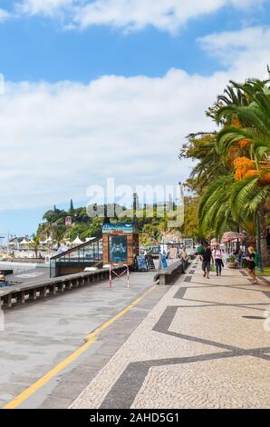 Funchal, Madeira, Portugal - Sep 10, 2019: City promenade along the harbor in the Madeiran capital. Cobbled pavement, green vegetation, palm tree and people on the streets. Vertical picture. Stock Photo