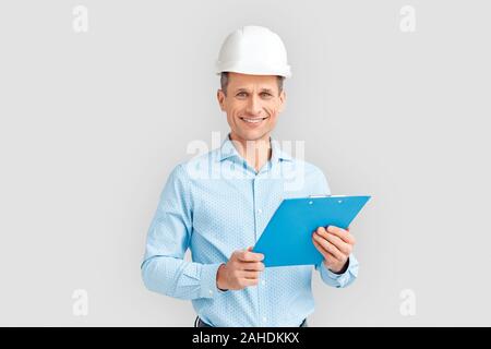 Mature man wearing hardhat studio standing isolated on white background holding clipboard looking camera smiling cheerful Stock Photo