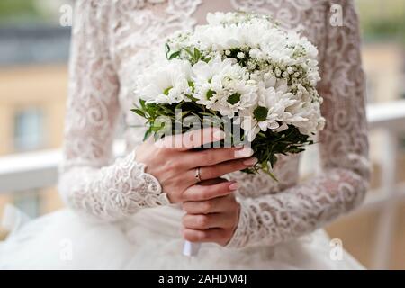 Bride in white lace dress is holding wedding flowers. Close-up on bride’s hands wearing engagement ring and holding white bouquet of flowers. Stock Photo