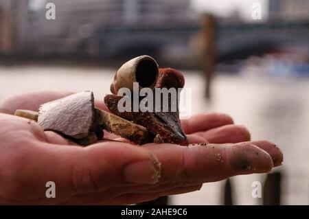 Mudlark findings: A sandy hand holds an old broken clay pipe and other pieces of the past found while mudlarking on the River Thames Stock Photo