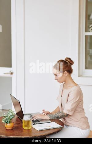 Concentrated young female freelancer sitting at small table at home and working on laptop Stock Photo