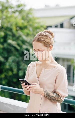 Beautiful young blond woman standing on balcony and using mobile application to make order online Stock Photo