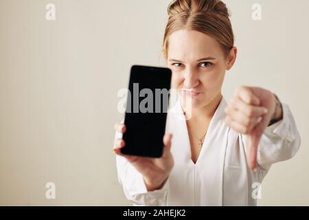 Frowning young businesswoman showing thumbs down when demonstrating smartphone with empty black screen Stock Photo