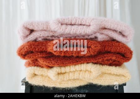 Three folded multi-colored sweaters lie on a stool in front of a curtain. Stock Photo
