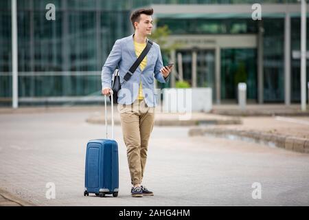 Stylish man standing at airport with smartphone and suitcase, browsing, texting, using mobile app. Business traveling. Stock Photo