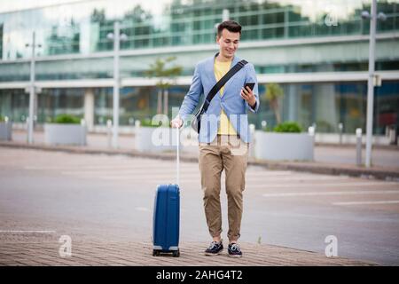 Stylish man standing at airport with smartphone and suitcase, browsing, texting, using mobile app. Business traveling. Stock Photo
