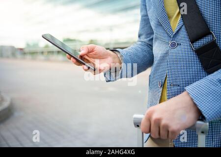 Stylish man standing at airport with smartphone and suitcase, browsing, texting, using mobile app. Businessman traveling. Close-up Stock Photo