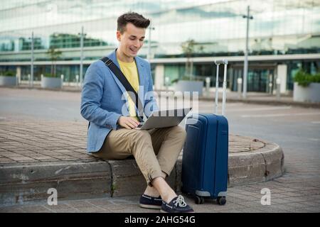 Stylish man sitting at airport with suitcase and laptop, working, typing, browsing. Businessman traveling. Stock Photo