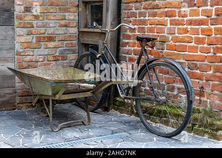 An old bicycle leaning against a red brick wall, next to a rusty wheelbarrow left at the entrance to the building. Stock Photo