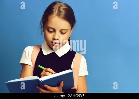 School girl with diligent face expression on blue background. Girl writes in big blue notebook with green pen, copy space. Pupil in school uniform with braid. Back to school and education concept Stock Photo