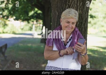 Young looking elder woman with smart phone leans on park tree. Blond senior lady text messaging on sunny morning outdoors. Communication, grandmother Stock Photo