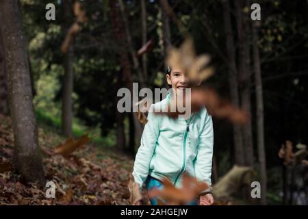 Smiling little girl watching brown leaves flying in forest. Playful kid with green fleece jacket having fun in woods on autumn day Stock Photo