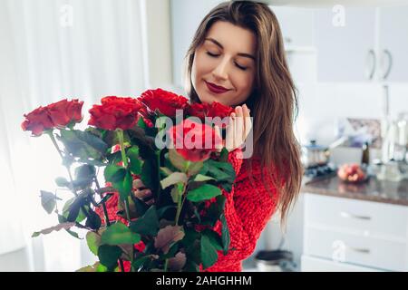 Valentines day. Young woman found bouquet of red roses on kitchen. Happy girl holding and smelling flowers. Stock Photo