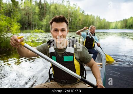 Two men in life vests canoeing in forest lake. Water surface covered with water lilies. Tourists traveling in Finland, having adventure. Stock Photo