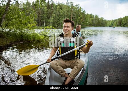 Two men in life vests canoeing in forest lake. Water surface covered with water lilies. Tourists traveling in Finland, having adventure. Stock Photo