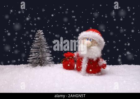 The toy knitted wool Santa Claus is standing next to a toy silver Christmas tree on artificial white snow against navy blue background Stock Photo