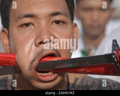 Young Thai Chinese Taoist devotee pierces his cheek with the blade and scabbard of an Asian sword during the Nine Emperor Gods Festival. Stock Photo