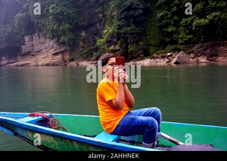 Close up of a boy taking a picture by his mobile phone sitting on a wooden boat in Dawki river, shillong wearing casual dress, selective focusing Stock Photo