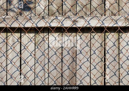 A weathered old gray wooden fence with a horizontal bar nailed by rusty nails stands behind a rusty metal mesh. Stock Photo