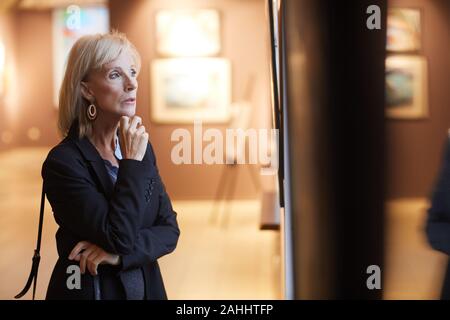Waist up portrait of elegant mature woman looking at paintings while enjoying exhibition in modern gallery or museum lit by dim light, copy space Stock Photo