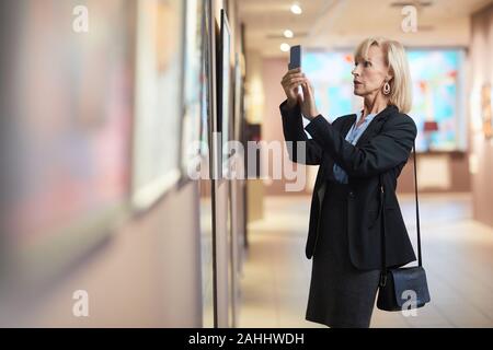 Side view portrait of elegant mature woman taking smartphone photo of painting while enjoying exhibition in art gallery or museum Stock Photo