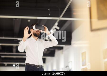 Waist up portrait of modern bearded man wearing VR gear while enjoying immersive experience during exhibition in art gallery, copy space Stock Photo