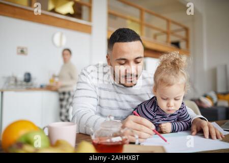 Portrait of loving mixed-race father drawing pictures with cute little girl while enjoying family time at home, copy space Stock Photo