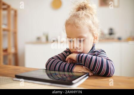 Warm-toned portrait of cute little girl looking at digital tablet while sitting at big table in cozy kitchen, copy space