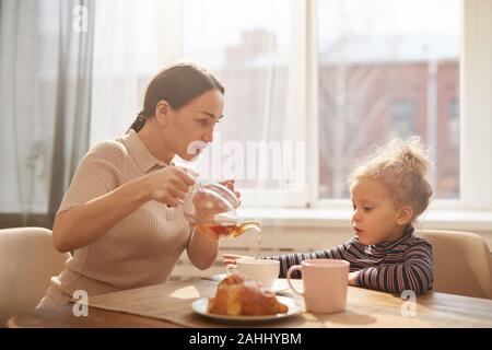 Side view portrait of modern mother pouring black tea for cute little girl while enjoying breakfast at home sitting against window in sunlight