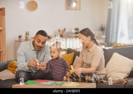 Warm toned portrait of happy mixed-race family playing with cute little daughter in cozy home interior
