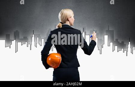 Businesswoman draws silhouette of new business center. Back view woman in business suit holding orange safety helmet. Architect presenting constructio Stock Photo