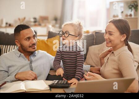 Portrait of happy young family counting home finances with cute little girl wearing glasses in sunlit apartment