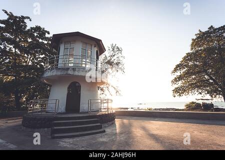 Old small lighthouse on the coast near street on the beach, with ray of sunset thru tree on backside Stock Photo