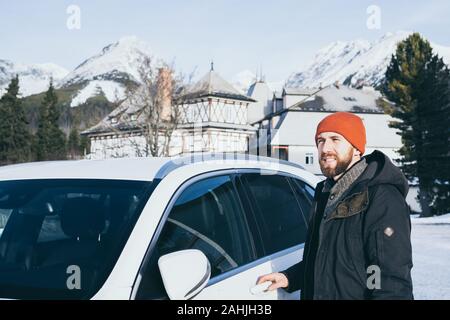 Bearded man standing next to white rental car in Strbske Pleso with High Tatras mountains on background, Slovakia. Stock Photo