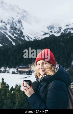 Woman having a hot drink from thermos cup overlooking snowcapped High Tatra mountains peaks Looking into the camera Stock Photo