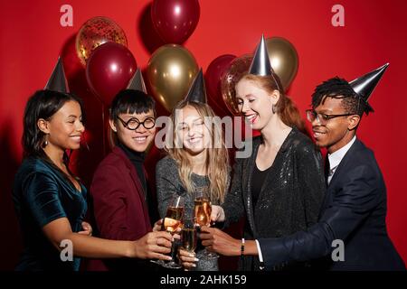 Multi-ethnic group of cheerful young people clinking champagne glasses while enjoying party standing over red background with balloons, copy space Stock Photo