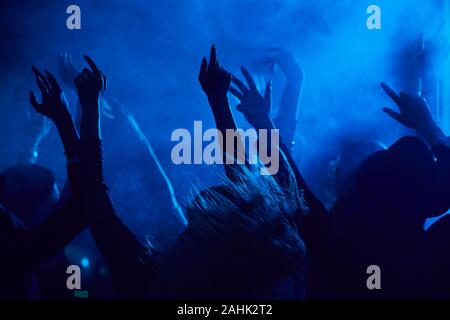 Silhouettes of young people jumping and raising hands while enjoying music concert in smoky nightclub lit by blue light, copy space Stock Photo