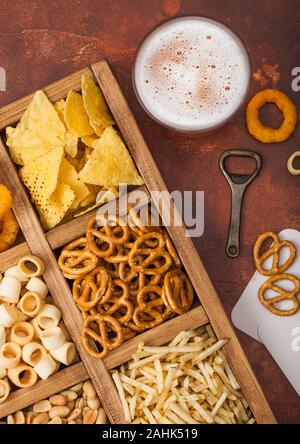 Glass of craft lager beer and opener with box of snacks on brown kitchen background. Pretzel,salty potato sticks, peanuts, onion rings with nachos in Stock Photo