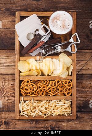 Glass of craft lager beer in vintage box of snacks openers and beer mats on wood background. Pretzel and crisps and salty potato. Stock Photo