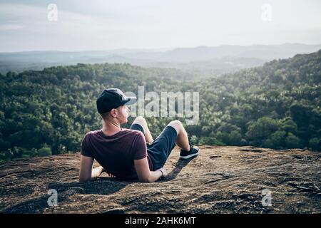 Pensive man resting on top of rock over jungle. Landscape with palm trees in Sri Lanka. Stock Photo