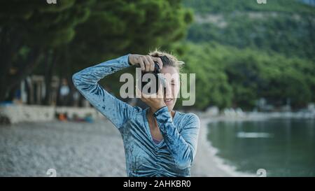 Front view of a young woman taking a photo directly at the camera standing outside on a pebble beach in early morning. Stock Photo