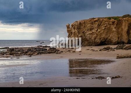 Coastline view of tall bluffs along El Matador Beach with reflections in the beach water and dramatic rain clouds in the distance, Malibu, California Stock Photo